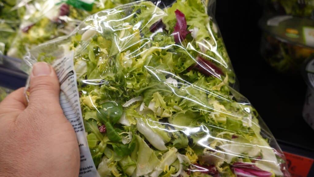A hand holds a sealed plastic bag of mixed salad greens in a grocery store produce section, checking the food expiration dates to ensure freshness.
