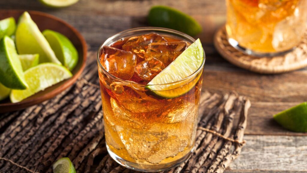 A glass of iced dark-colored cocktail crafted by expert rum distillers is garnished with a lime wedge on a rustic wooden surface, with lime slices in a bowl and more limes in the background.