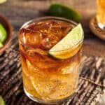 A glass of iced dark-colored cocktail crafted by expert rum distillers is garnished with a lime wedge on a rustic wooden surface, with lime slices in a bowl and more limes in the background.