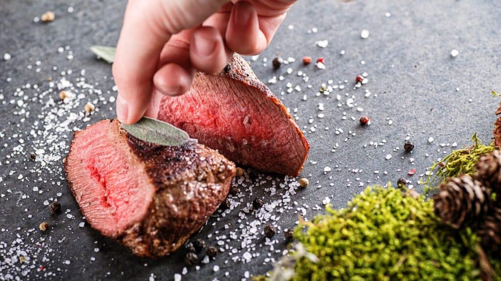 A hand places a sage leaf on two slices of rare-cooked steak—foods facing bans or restrictions in some regions—sprinkled with coarse salt and pepper, on a dark slate surface with moss and pine cones nearby.