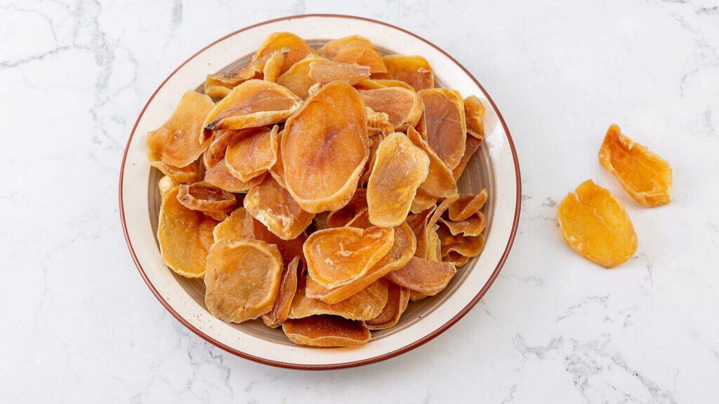 A plate filled with dried mango slices, one of the many foods shaped by US military innovations, sits on a white marble surface, with a few slices scattered beside the plate.