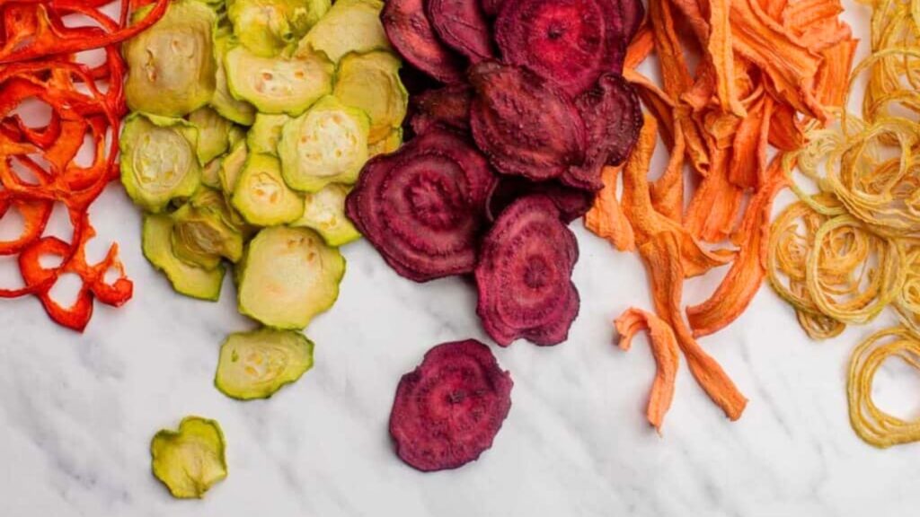 Assorted dried vegetable chips, including red, green, purple, orange, and yellow varieties&mdash;some of the best foods to dehydrate&mdash;arranged in small piles on a white marble surface.