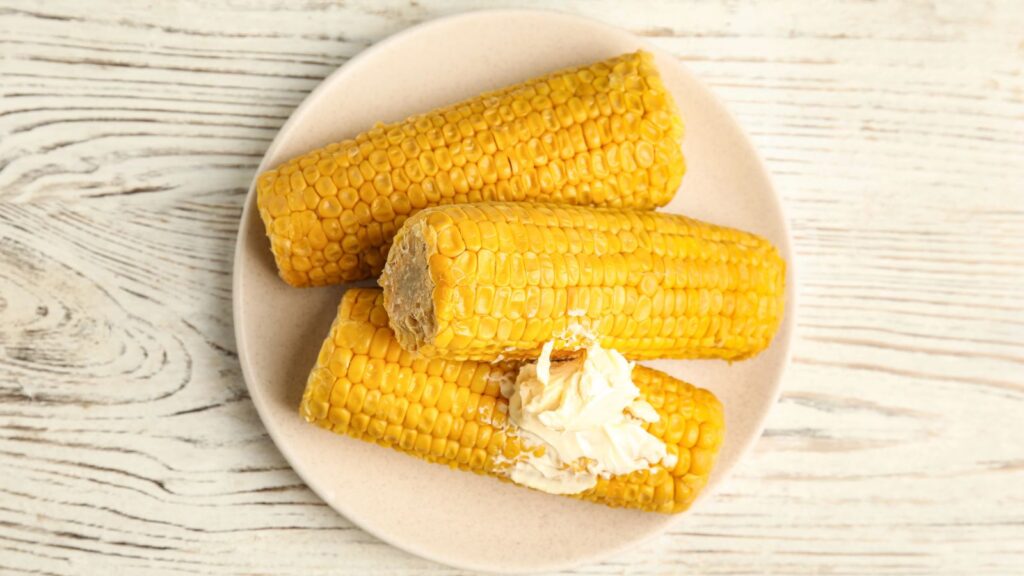 Three cooked corn on the cob pieces on a beige plate, with one piece topped with a portion of butter.