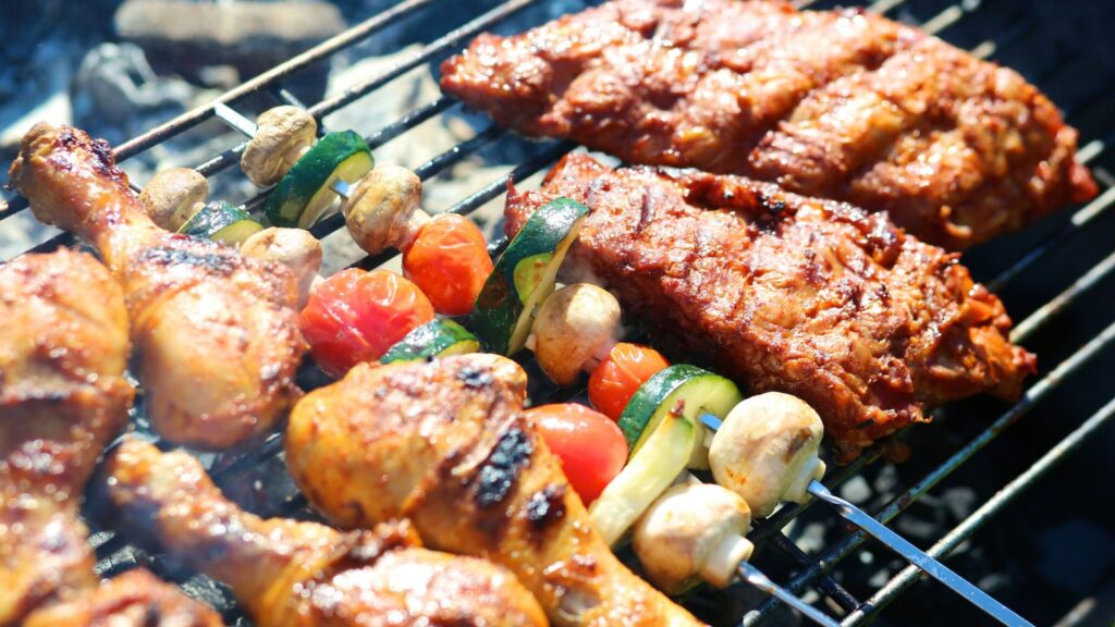 Grilled chicken drumsticks, skewers with vegetables, and ribs cooking on an outdoor barbecue grill.
