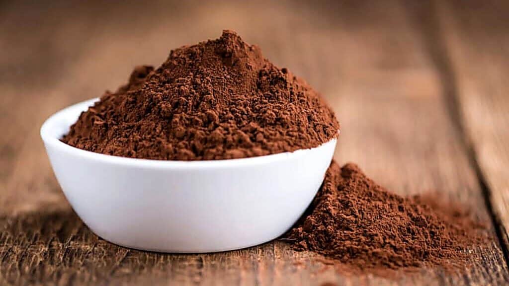 A white bowl filled with cocoa powder sits on a wooden surface, with some powder spilled beside it.