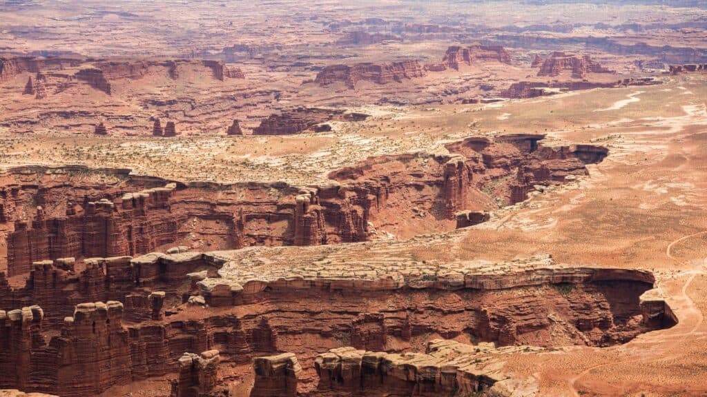 Aerial view of deep, layered red rock canyons and mesas in a desert landscape under a hazy sky, reminiscent of iconic scenery found in US national parks where reservations are now often required.