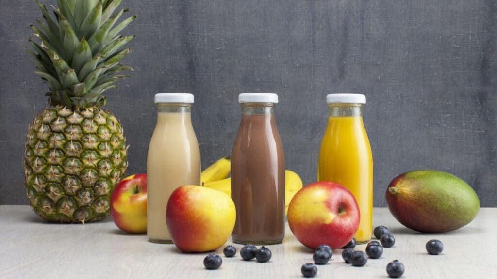 Three glass bottles with white caps contain beige, brown, and yellow liquids, surrounded by a pineapple, apples, bananas, a mango, and blueberries on a light surface&mdash;a vibrant scene reminding us that food labeling terms aren&rsquo;t simple.