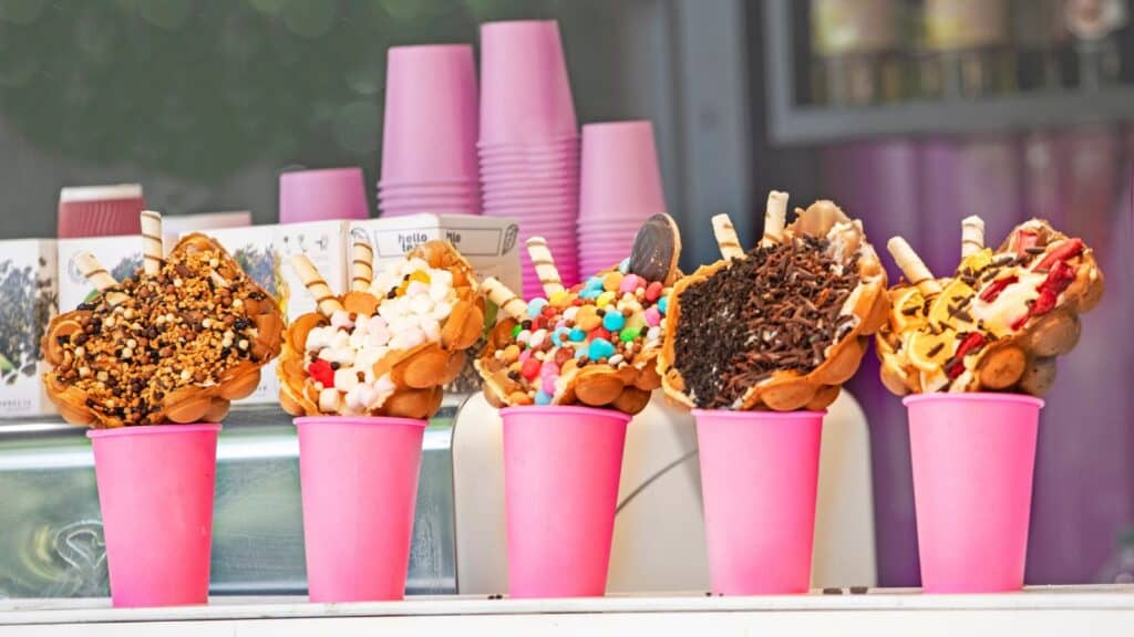 Five pink cups hold bubble waffles topped with various ice creams, candies, chocolate, and wafers, displayed in a row next to stacked pink cups.