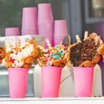 Five pink cups hold bubble waffles topped with various ice creams, candies, chocolate, and wafers, displayed in a row next to stacked pink cups.