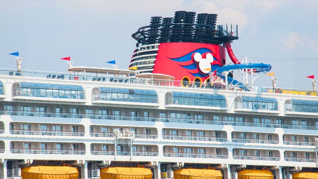 Close-up of a Disney Cruise Line ship featuring a red funnel with a Mickey Mouse logo, slide, lifeboats, and balconies.