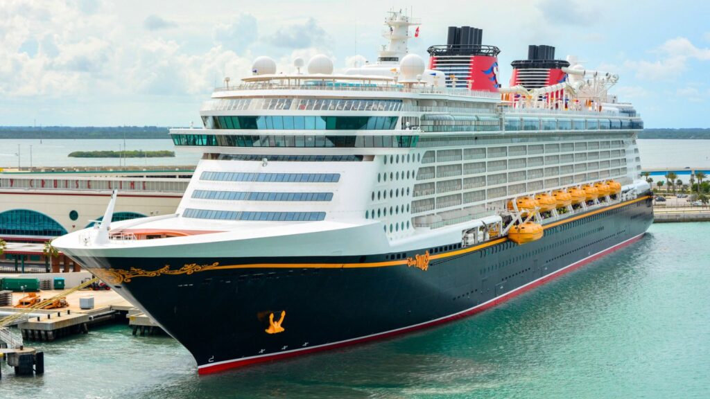 Large cruise ship docked at a port, with lifeboats along the side and two red-and-black funnels on top; the sky is partly cloudy.
