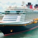 Large cruise ship docked at a port, with lifeboats along the side and two red-and-black funnels on top; the sky is partly cloudy.