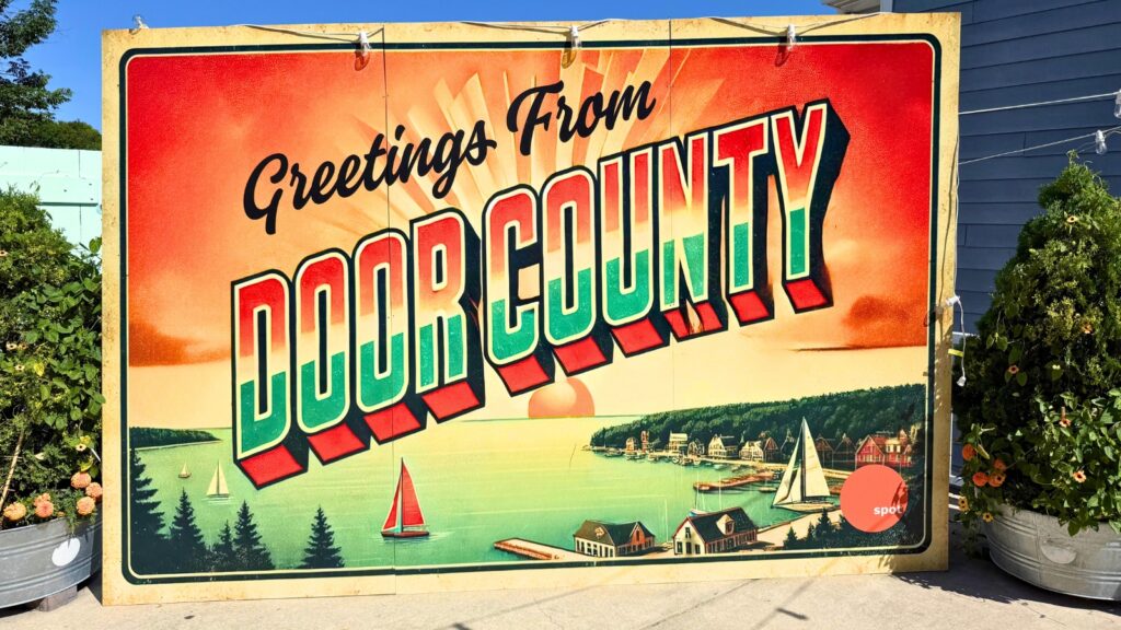 Large vintage-style sign reads “Greetings From Door County” with an illustrated lake scene, sailboats, and cottages at sunset.