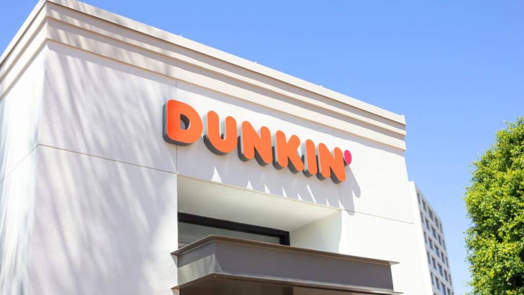 A white Dunkin’ building with an orange and pink logo on the exterior, viewed from below against a clear blue sky.