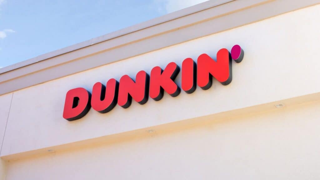 The exterior of a Dunkin’ store features a large red and pink “DUNKIN’” sign on a cream-colored wall.
