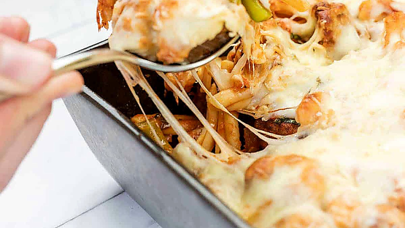 A hand uses a spoon to serve Easy Cheesy Meatball Casserole from a rectangular baking dish, with melted cheese stretching from the dish.