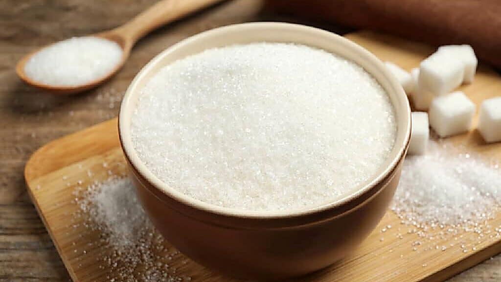 A bowl filled with granulated white sugar sits on a wooden board, with sugar cubes and a wooden spoon in the background.