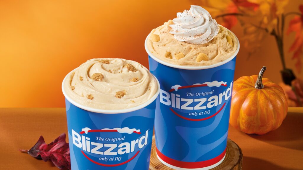 Two Dairy Queen Blizzard cups filled with creamy desserts, including a Pumpkin Pie Blizzard topped with whipped cream and cinnamon, set on a wooden surface with autumn decorations and a small pumpkin in the background.