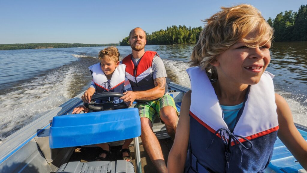 Three people wearing life jackets ride in a small motorboat on a lake, experiencing the thrill of America&rsquo;s boating tradition, with trees in the background and water trailing behind them.