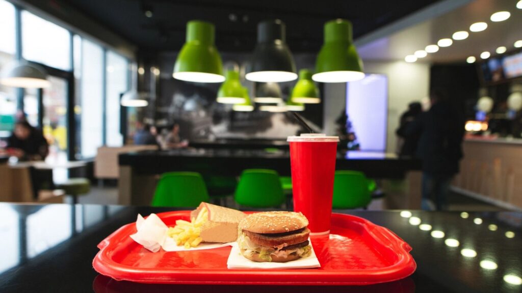 A red tray with a burger, fries, and a drink sits on a black table in a modern fast-food restaurant with green chairs and hanging lights.