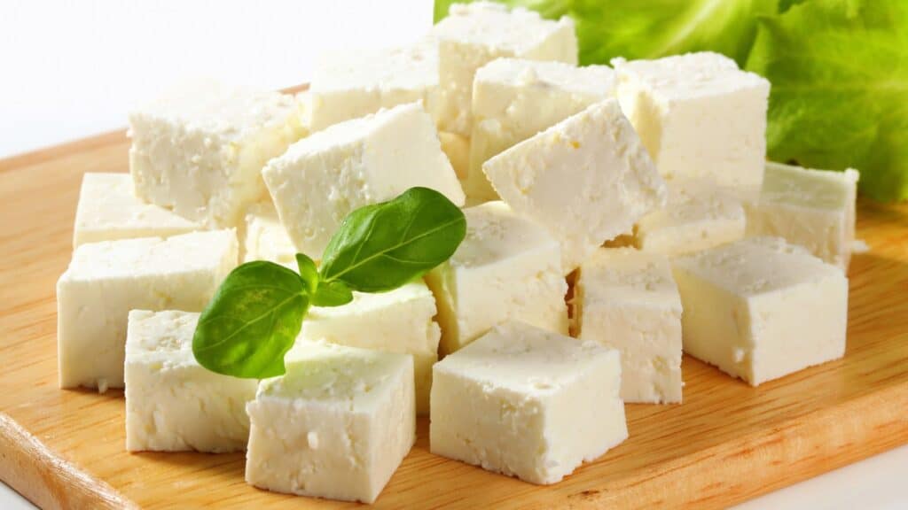 Cubes of white cheese arranged on a wooden board with a sprig of green basil and lettuce leaves in the background, showcasing one of the many delicious foods with protected names in Europe.