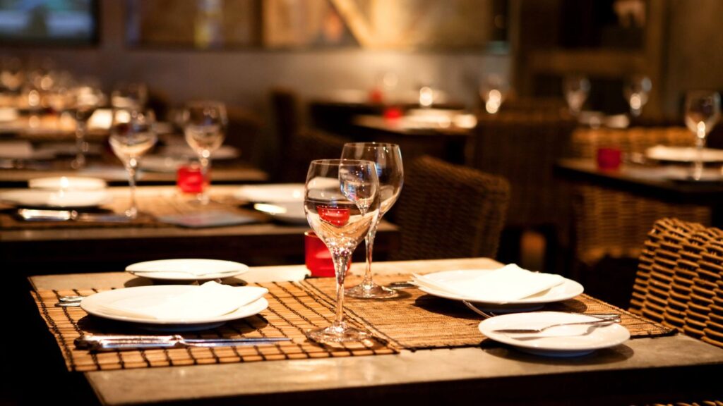 A restaurant table set with plates, cutlery, and empty wine glasses on a woven placemat, with more tables and chairs in the background.
