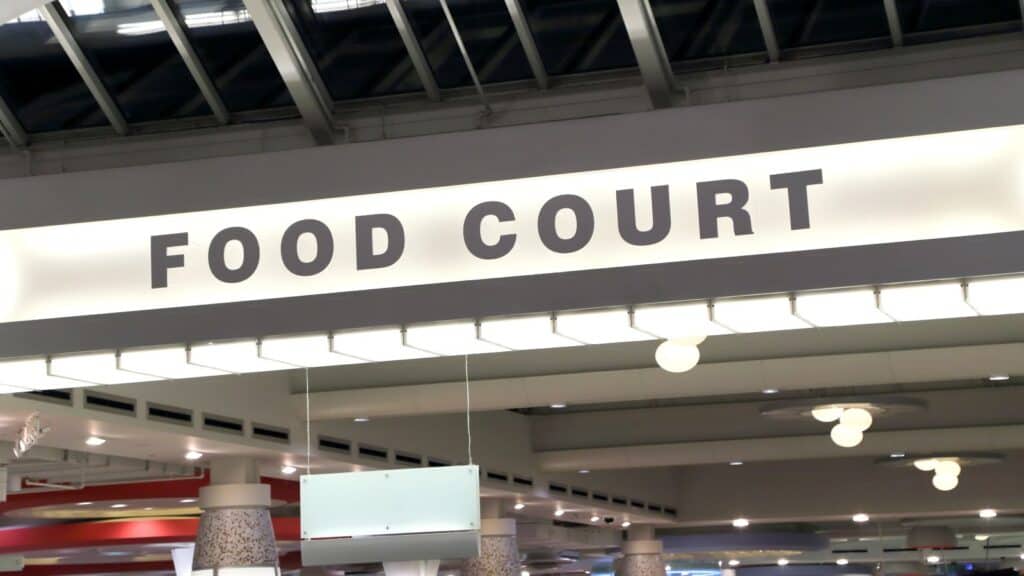 A white sign with bold black letters reading “FOOD COURT” hangs indoors under a skylight at a shopping mall or public space, inviting visitors to enjoy classic mall food court staples.