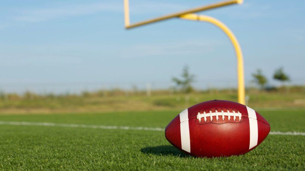 A football rests on a grassy field near a yellow goalpost under a clear sky.