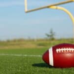 A football rests on a grassy field near a yellow goalpost under a clear sky.