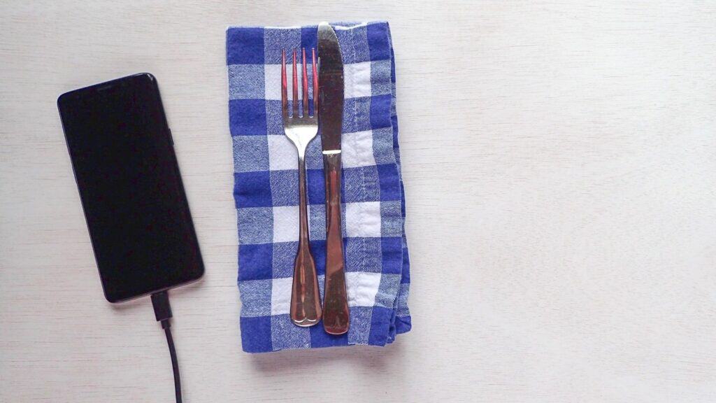 A fork and knife rest on a blue plaid napkin next to a charging smartphone on a light-colored table—a simple setup perfect for those looking to get work done in airport lounges or cafés.