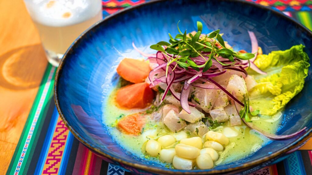 A blue bowl with ceviche, featuring chunks of fish, red onion, corn, greens, and sweet potato, on a colorful patterned tablecloth. A drink with foam is in the background.