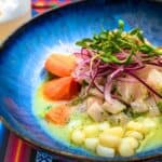 A blue bowl with ceviche, featuring chunks of fish, red onion, corn, greens, and sweet potato, on a colorful patterned tablecloth. A drink with foam is in the background.