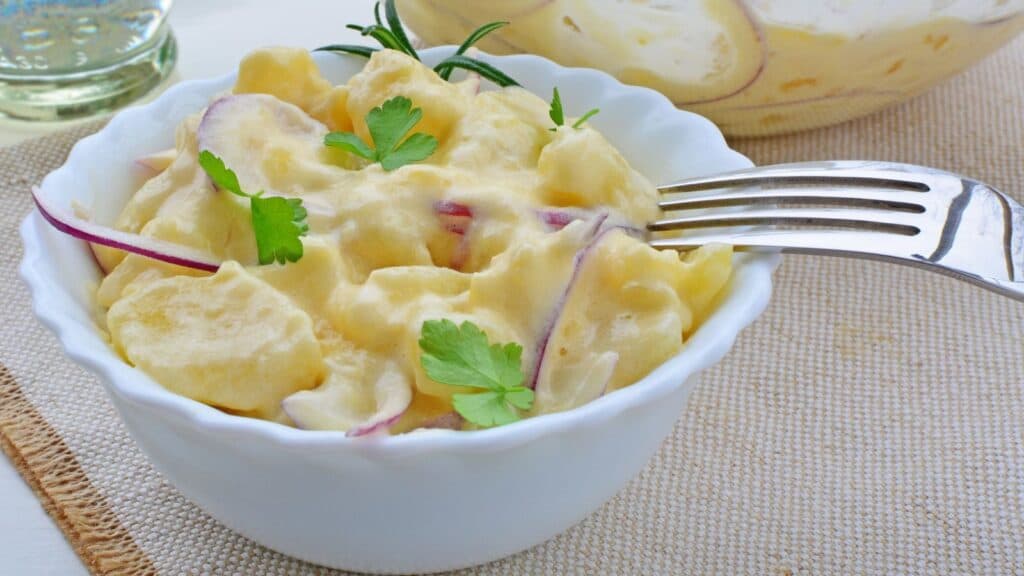 A white bowl filled with creamy potato salad&mdash;a classic example of American foods with foreign roots&mdash;is garnished with parsley and thinly sliced red onion, with a fork resting on the bowl.