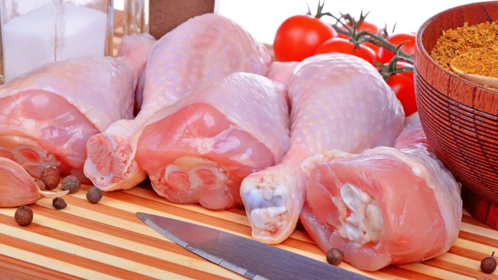 Raw chicken drumsticks on a wooden cutting board with whole peppercorns, a knife, tomatoes, a spice blend, and a salt shaker nearby&mdash;a simple setup reflecting how Americans eat compared to the stricter EU rules on food preparation.