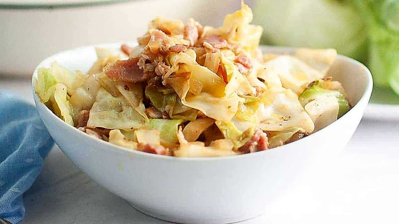 A white bowl filled with fried cabbage with pieces of bacon sits on a white surface with a blue cloth nearby.