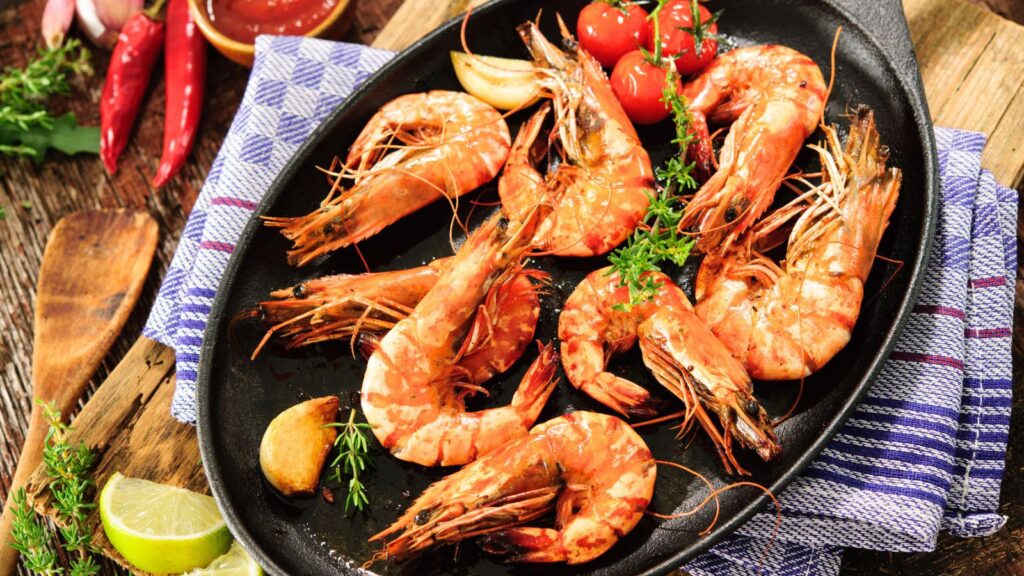 Cooked shrimp with herbs, cherry tomatoes, and garlic arranged on a black plate, with a lime wedge, chili peppers, and a checkered napkin visible nearby.