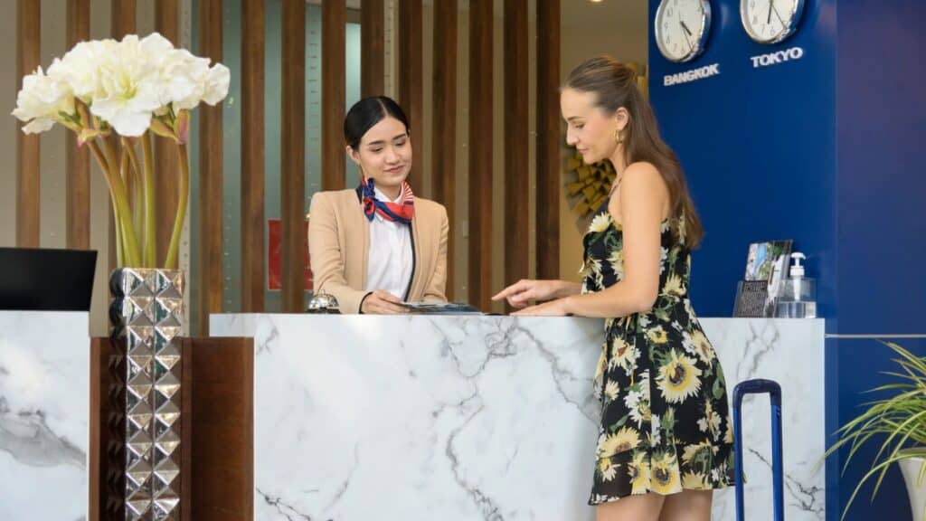 Two women stand at a hotel reception desk; one is a receptionist in uniform&mdash;often one of the hotel concierges guests turn to for assistance&mdash;and the other is a guest with a suitcase. Clocks showing different time zones are on the wall behind them.