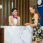Two women stand at a hotel reception desk; one is a receptionist in uniform—often one of the hotel concierges guests turn to for assistance—and the other is a guest with a suitcase. Clocks showing different time zones are on the wall behind them.