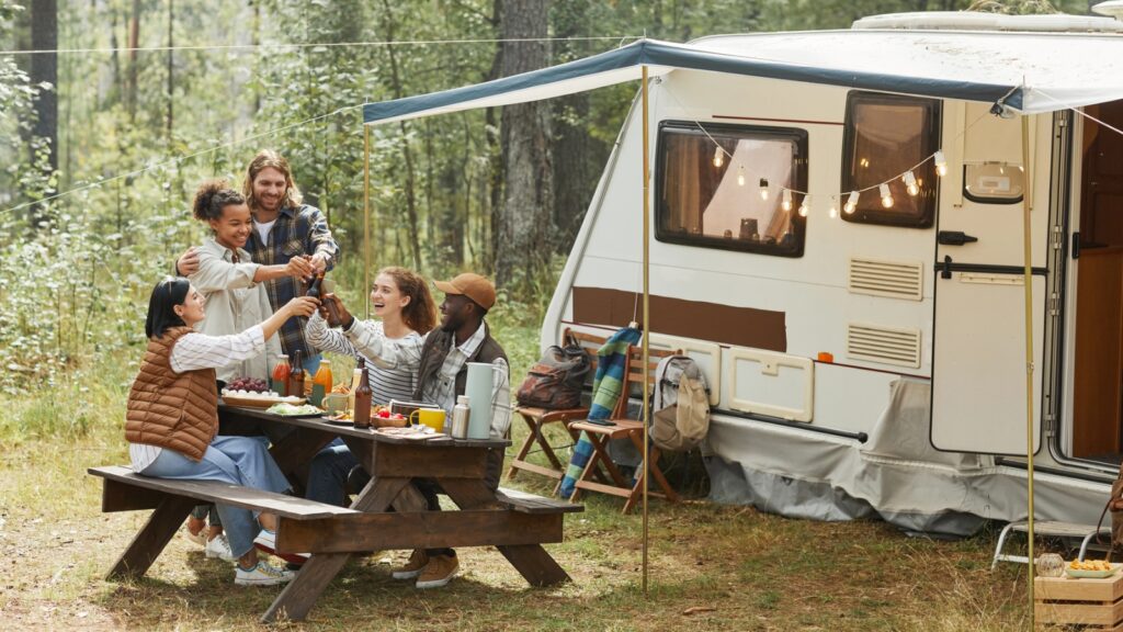 Five people sit at a picnic table sharing drinks and food next to a camper in a wooded area, with string lights hanging above.