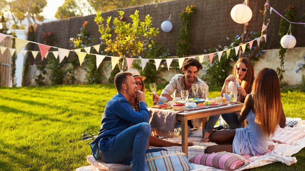 Five people sit on blankets around a low table having food and drinks at an outdoor picnic in a sunny backyard decorated with bunting and lanterns.