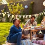 Five people sit on blankets around a low table having food and drinks at an outdoor picnic in a sunny backyard decorated with bunting and lanterns.