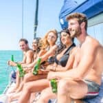 Five young adults sit on a sailboat in swimwear, holding beer bottles, smiling and looking toward the water on a sunny day.