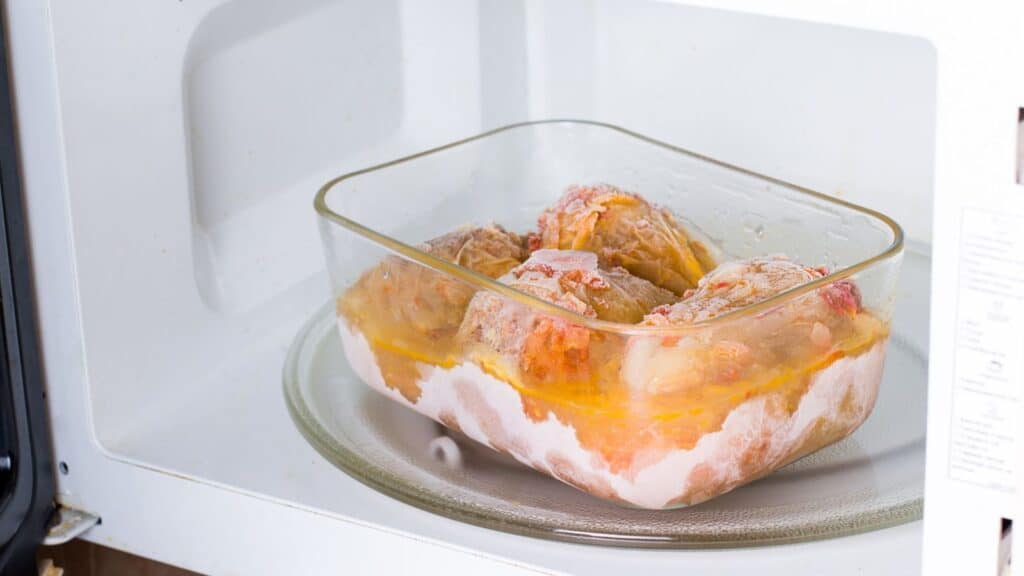 A glass dish with a partially thawed frozen dinner, shaped by the convenience of the American freezer, sits on the rotating plate inside a microwave oven.