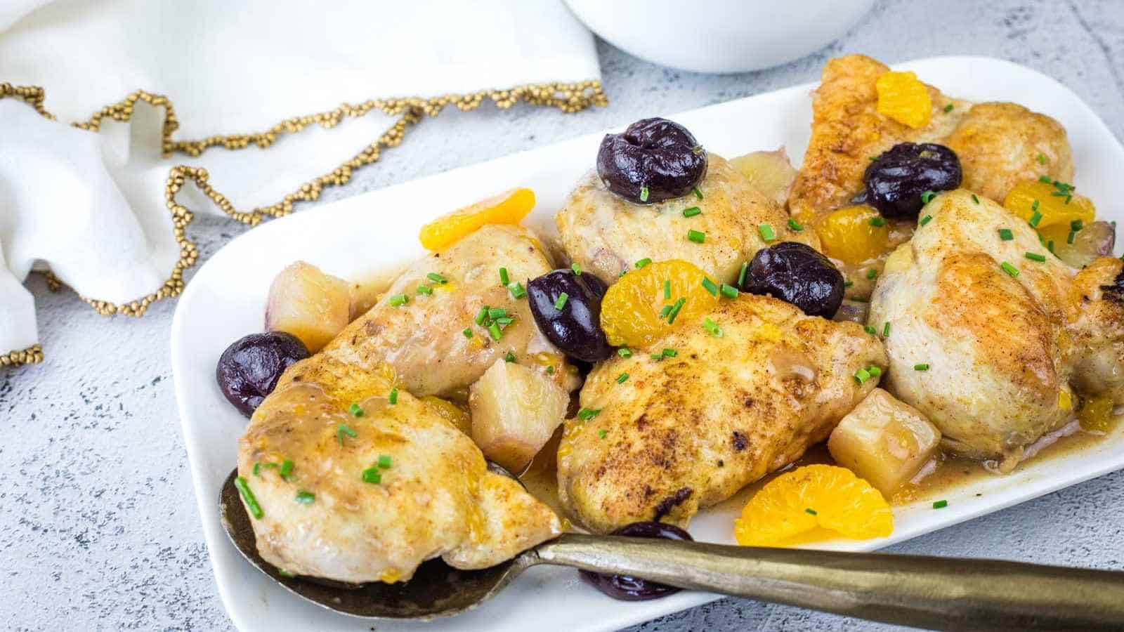 Fruited chicken pieces garnished with chopped herbs, served with cherries, orange slices, and pineapple chunks on a white rectangular plate. A serving spoon rests on the dish.