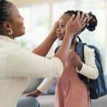 An adult adjusts a young girl&rsquo;s hair as she wears a backpack and stands in a living room, preparing for what appears to be a school day.