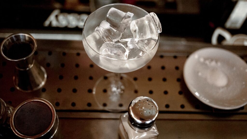 A cocktail glass filled with ice cubes sits on a bar counter next to a salt shaker&mdash;a classic example of things Americans ask for abroad&mdash;accompanied by a small plate and bartending tools.