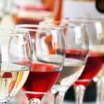 Several glasses of white and red wine are arranged closely together in rows, with bottles visible blurred in the background.
