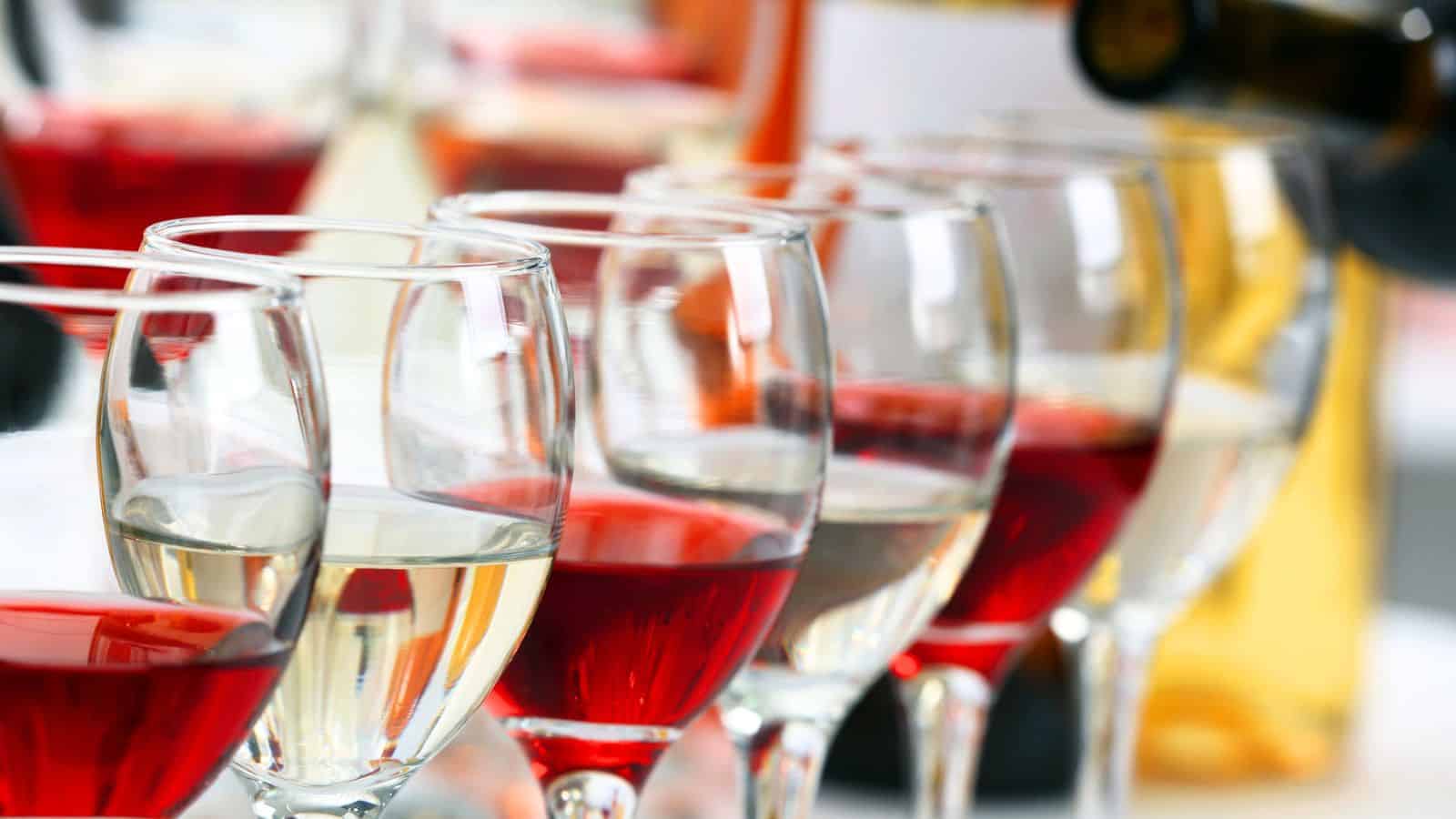 Several glasses of white and red wine are arranged closely together in rows, with bottles visible blurred in the background.