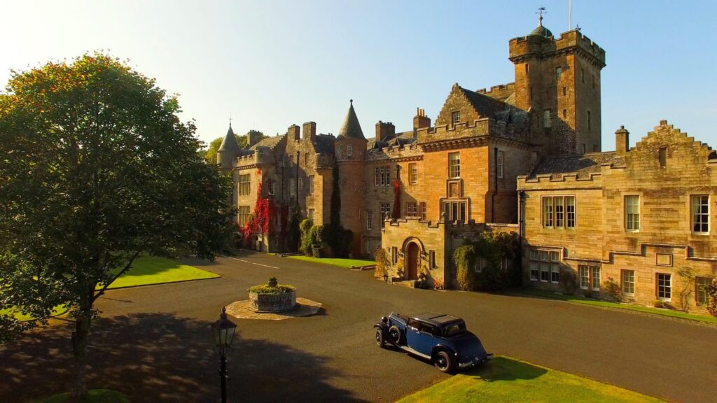 A historic stone castle with turrets and ivy-covered walls is seen from above, with a vintage blue car parked on the driveway and green lawns surrounding the property.