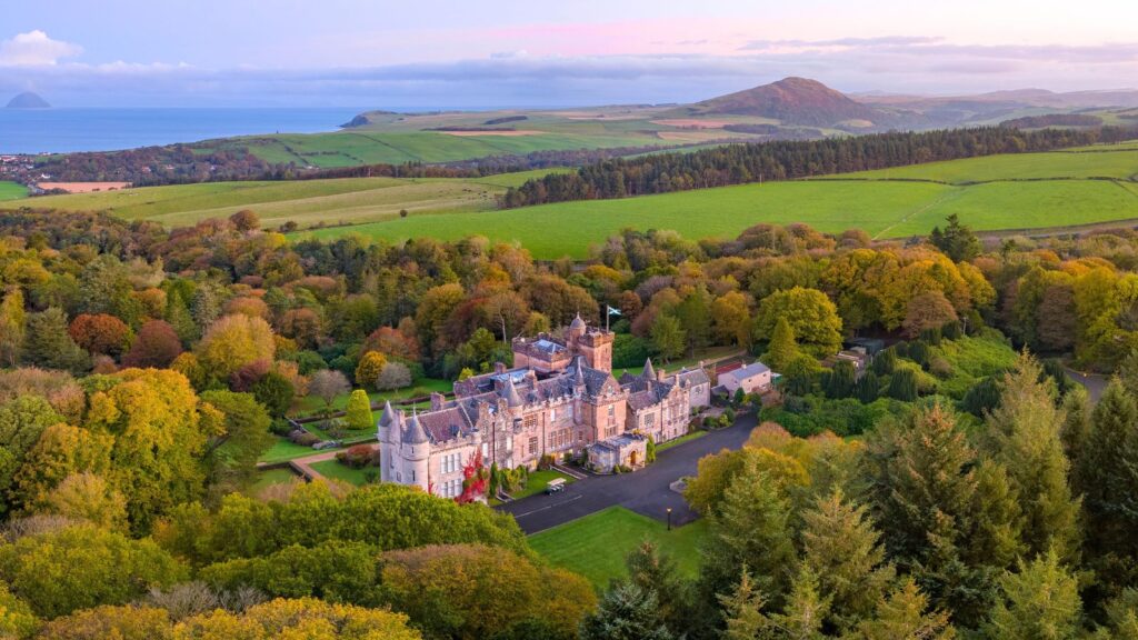 A large stone castle with multiple towers sits among dense green trees, with rolling hills and the ocean visible in the background under a pastel sky.
